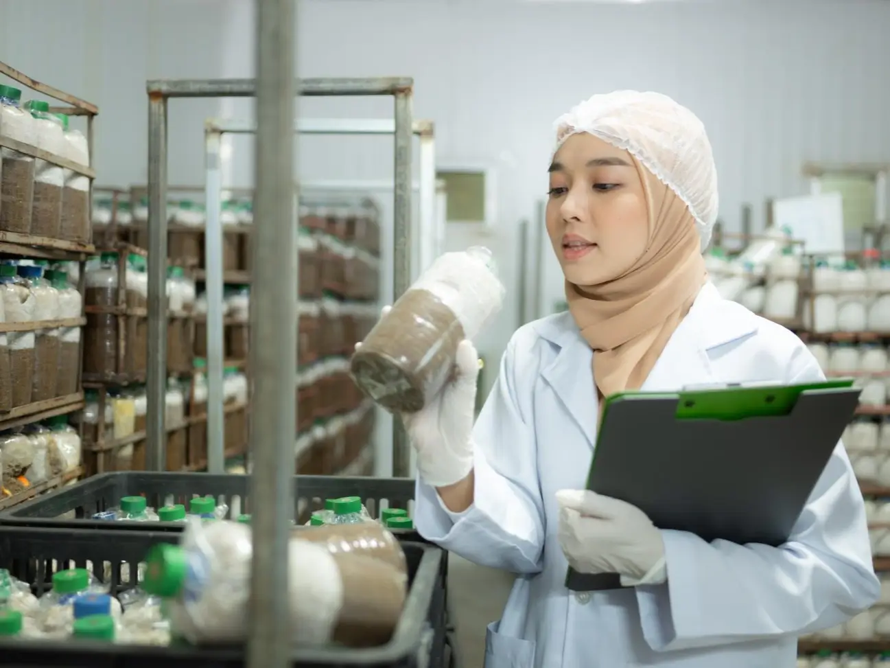 Food safety technician inspecting containers in a food production facility, wearing a lab coat, gloves, and a headscarf, with bottles of ingredients or products stacked in the background